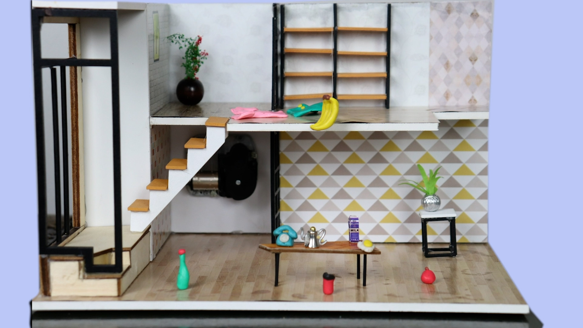 photo of a miniature house placed on a periwinkle background. Clay-sculpted items are strewn around the house: upstairs, a shirt and skirt with large buttons. Downstairs: a green water bottle, a travel mug, an apple, and a plant. A rotary telephone, milk carton, egg and tea kettle sit on a table in the centre. Bananas are being lowered from the top level, hanging by a fishing wire.